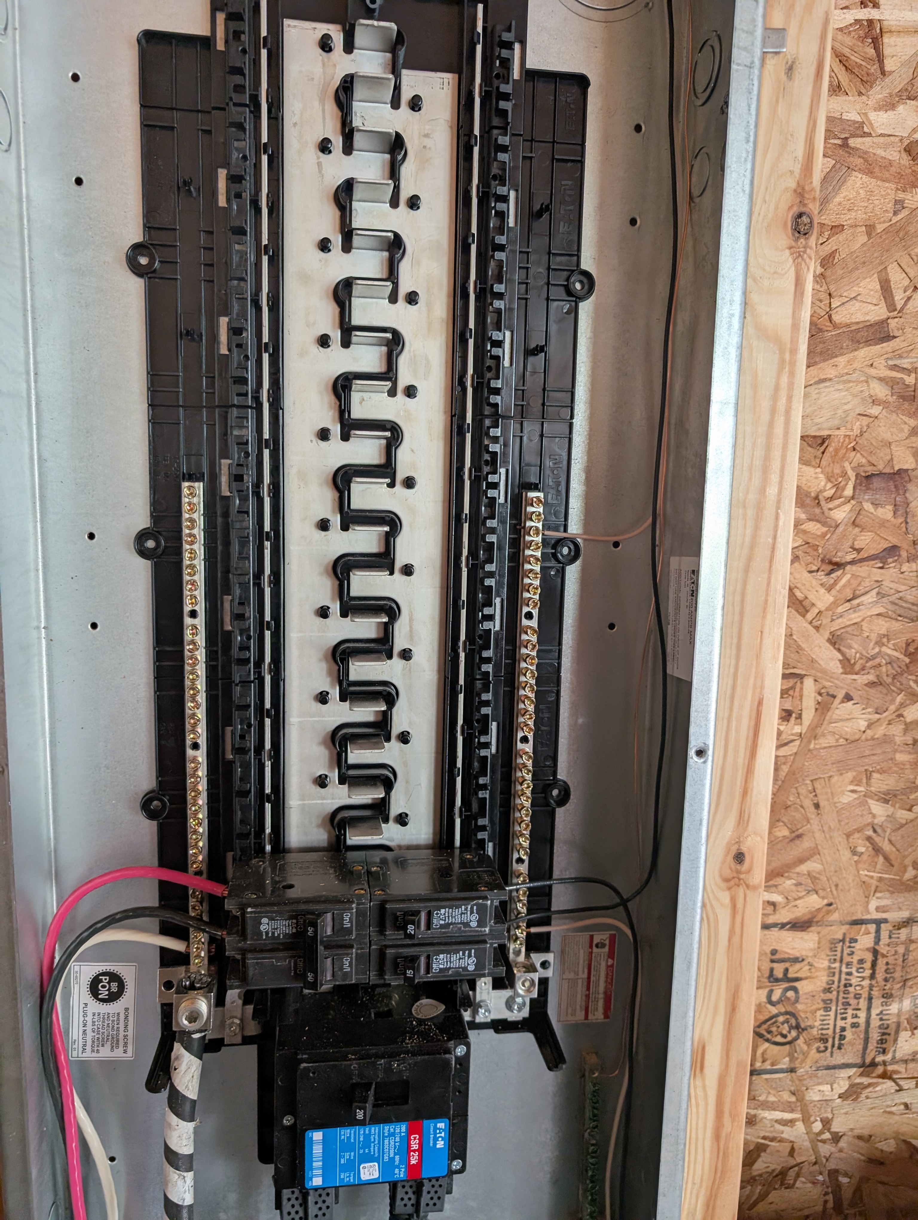 Eaton 200A garage panel with the dead-front cover removed; empty breaker rails / bus stabs visible above; main lug compartment at bottom with feeder cables and two 2-pole branch breakers landed (SLS-installed circuits — interior GFCI + soffit lights / exterior GFCI); panel mounted on OSB-sheathed stud wall