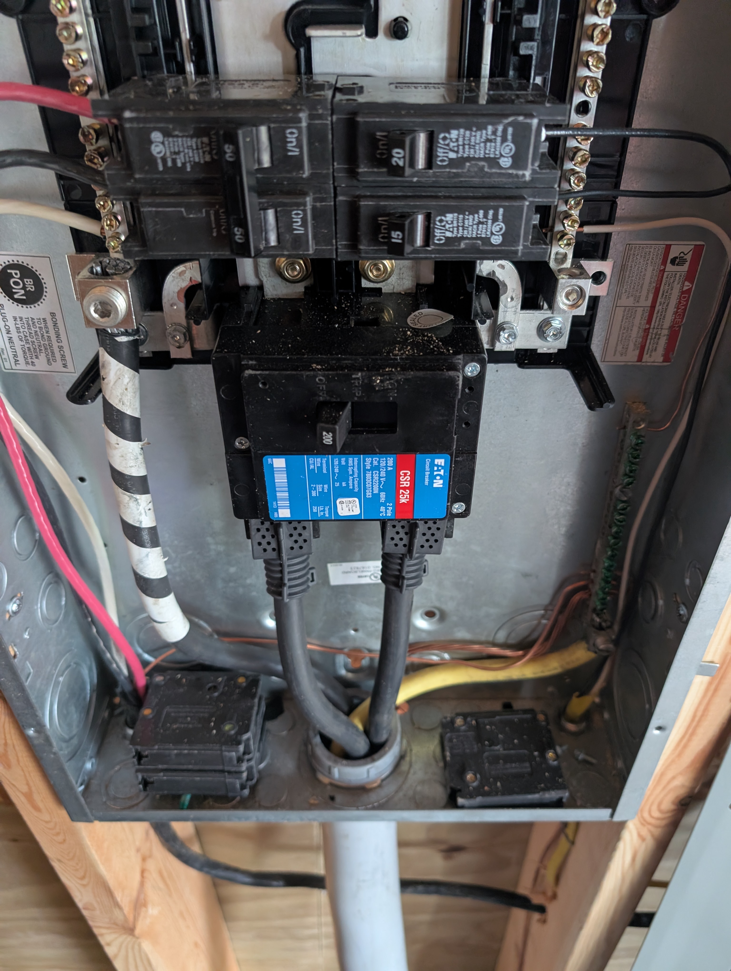 Close-up of the bottom of the garage Eaton panel — large blue-labeled CSR 200A main breaker at center; four populated branch breaker positions above (SLS-installed circuits); main feeder cables routed into the lugs; two 1" conduit knockouts at the bottom for low-voltage runs