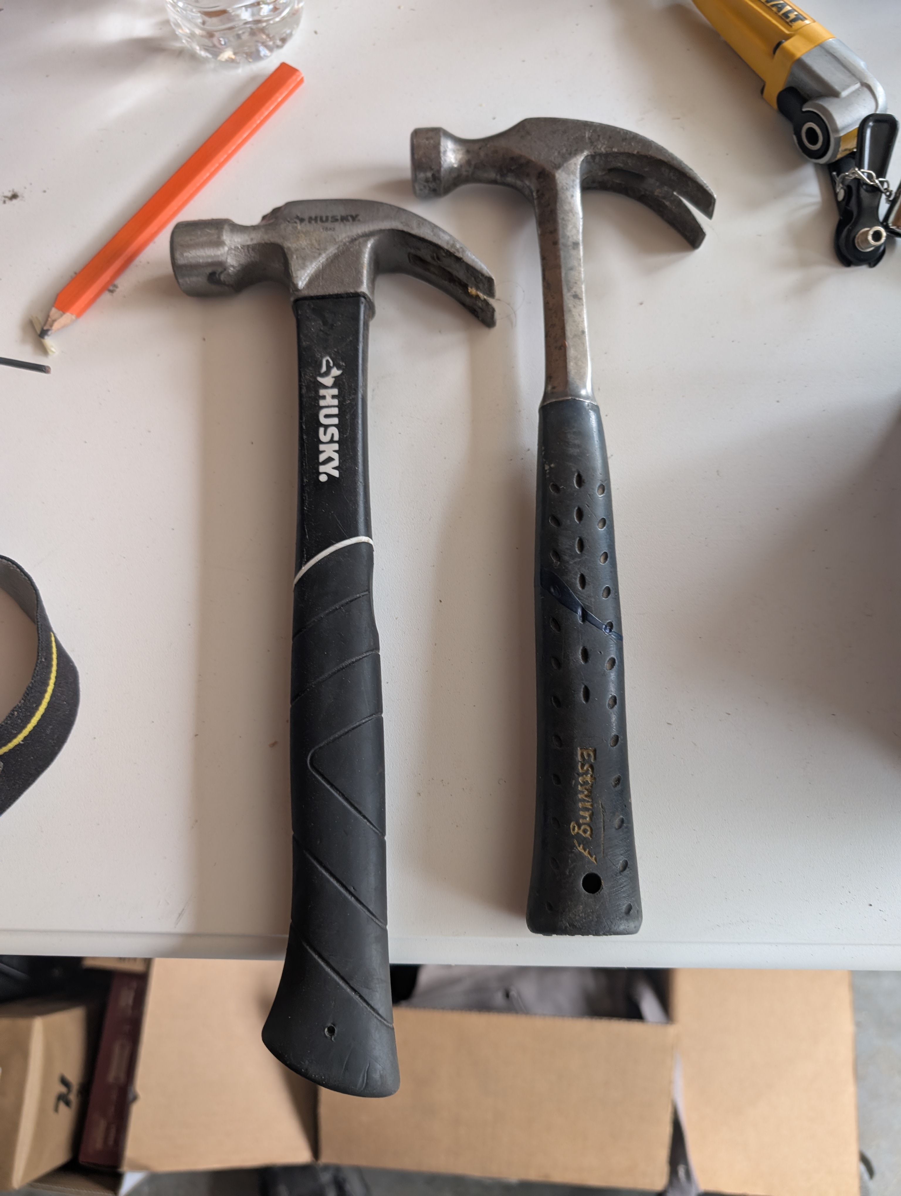Two claw hammers on a white tabletop — left: Husky claw hammer with black rubber-overmolded handle; right: Estwing one-piece steel claw hammer with worn rubber grip showing heavy use and decades of patina