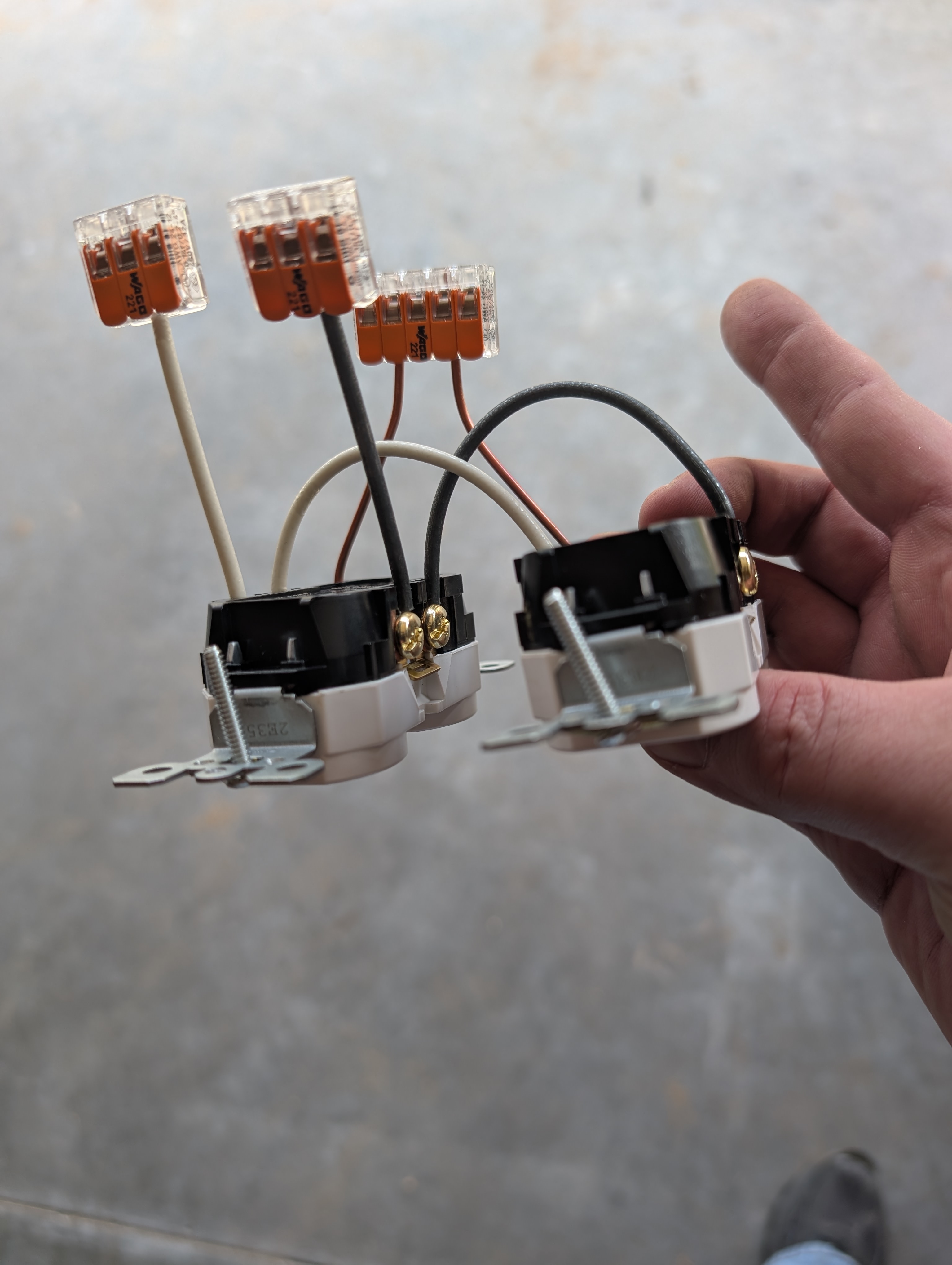 2-gang outlet pair held by hand, with three WAGO 221 lever-nuts stacked above the assembly — long pigtails extending up from each WAGO ready to land on the incoming and outgoing 12/2 NM-B in the box