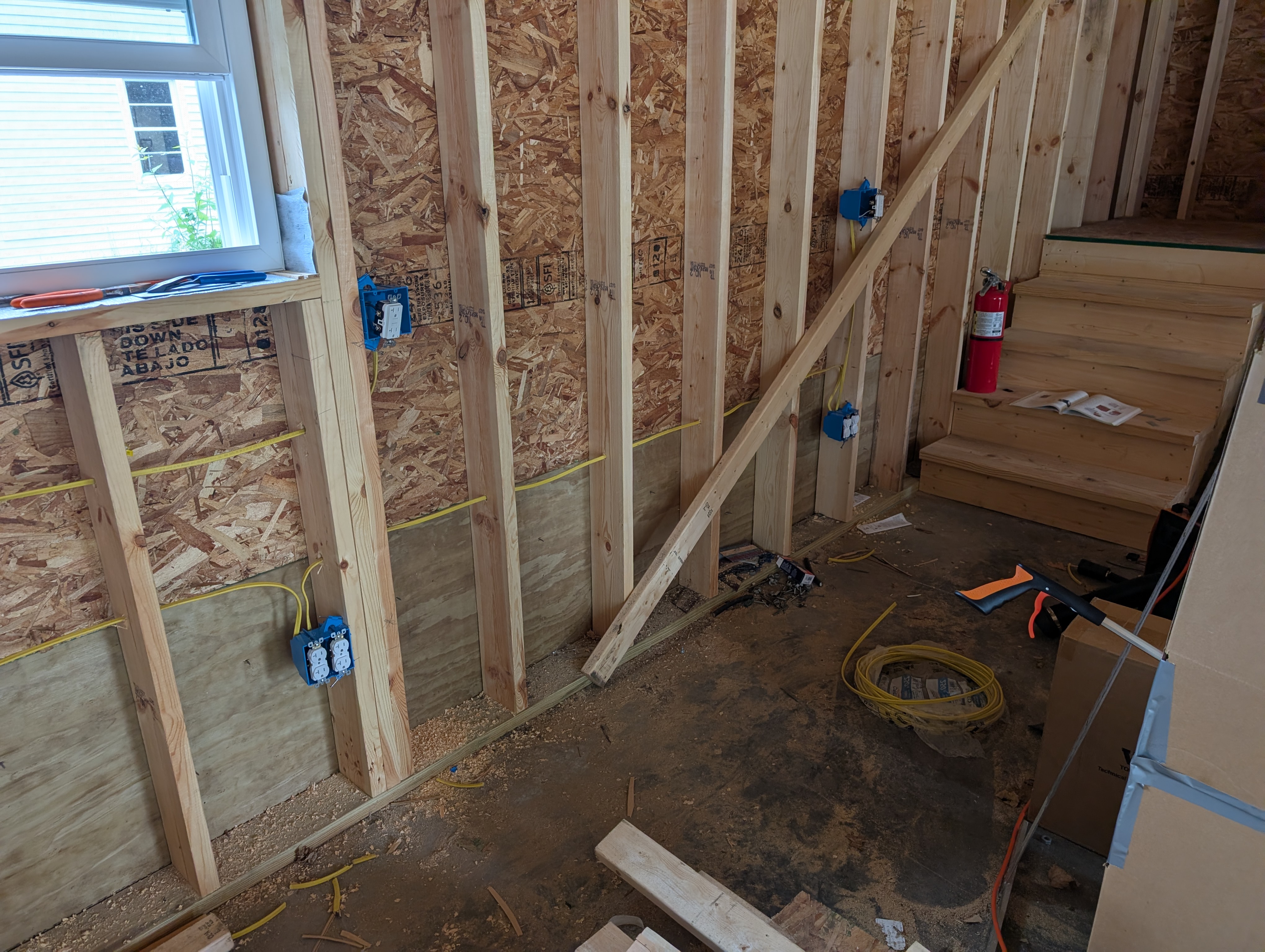Wide view of the rear (south) garage wall — three blue 2-gang plastic boxes mounted at 16" AFF, one workbench-height box visible higher on the same wall, plywood lower wall protection, stairs and red fire extinguisher at right