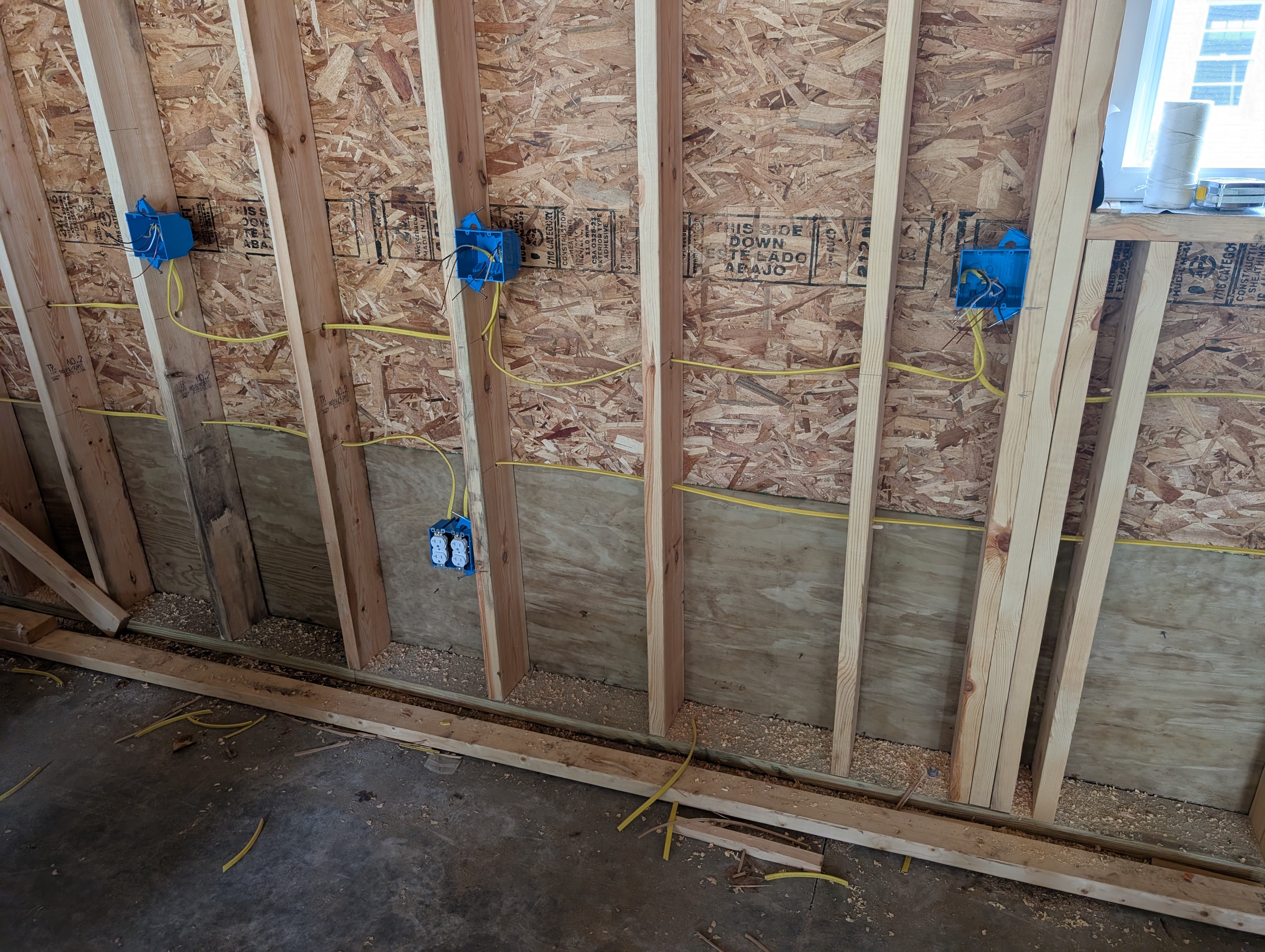 Closer view of the rear wall outlet run — four 2-gang plastic boxes at 16" AFF chained with yellow 12-2 NM-B romex visible between boxes; leftmost box holds the GFCI device protecting the downstream chain; one workbench-height box at far right