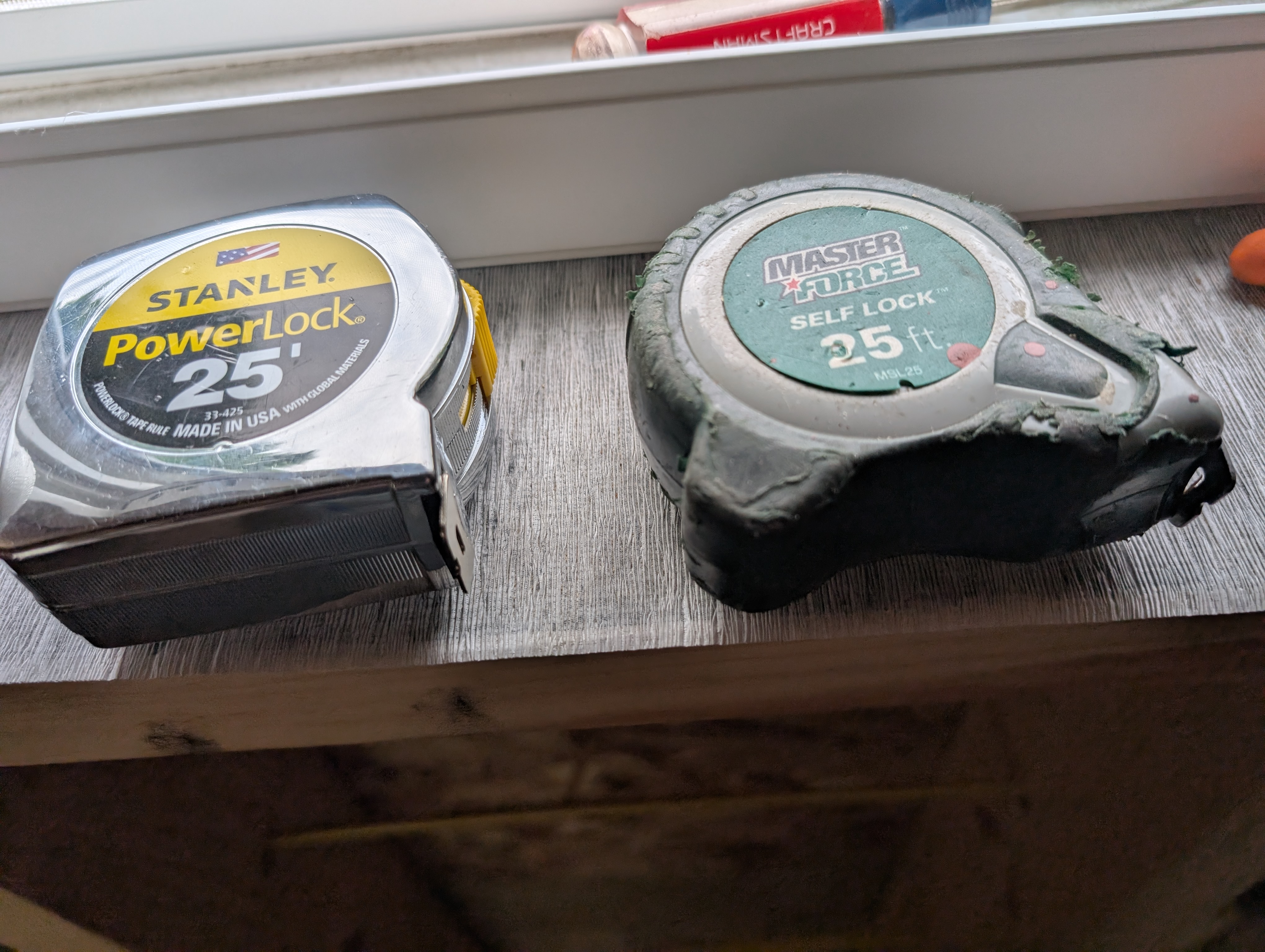 Two 25' tape measures resting on a windowsill — left: clean Stanley PowerLock with chrome housing and yellow label; right: Master Force Self Lock with green paint/mud splattered all over the casing, decades of use