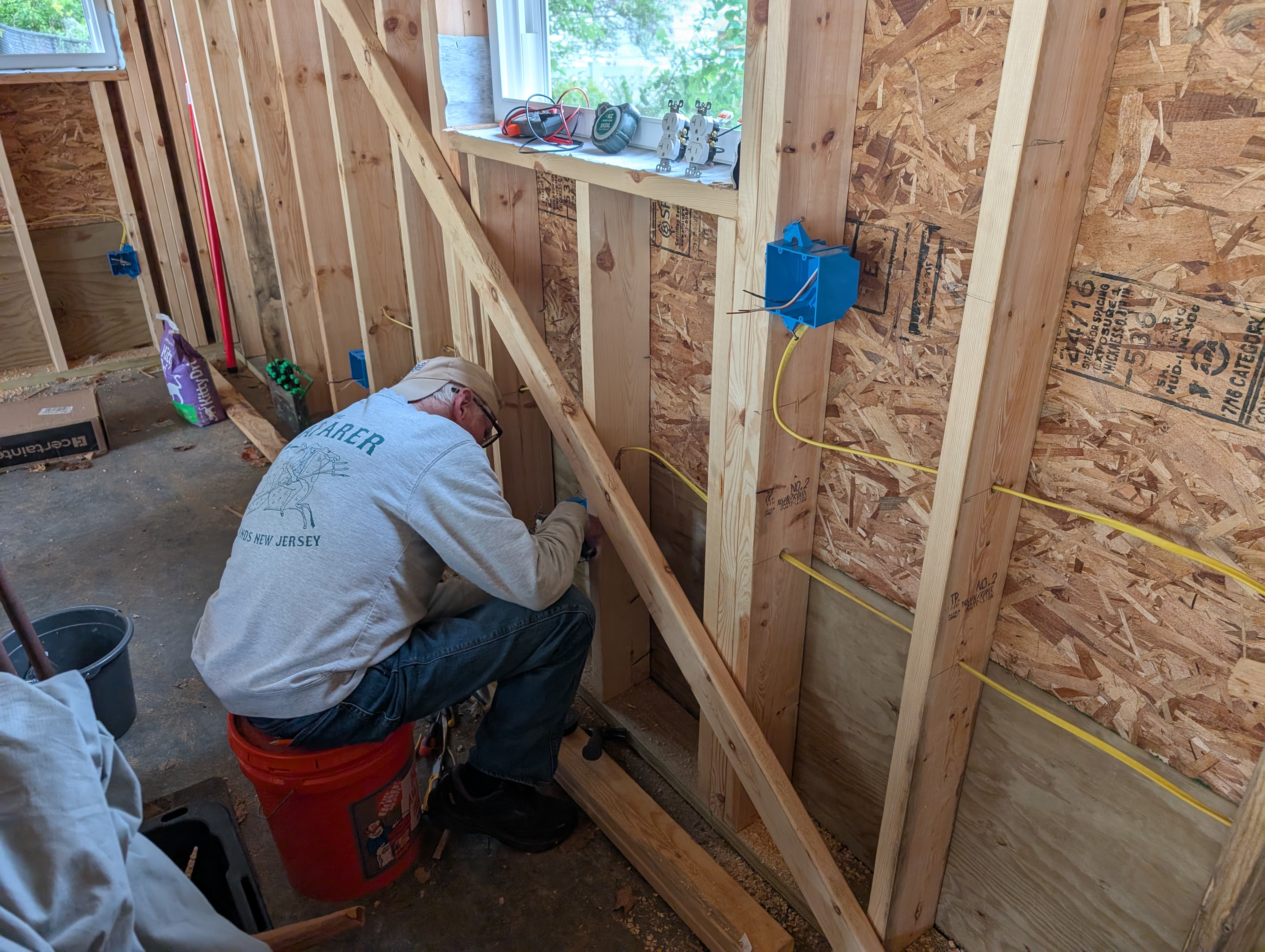 Uncle Ed kneeling on a flipped 5-gallon bucket, working low at the north/west corner of the garage; to the right the north wall runs off with a workbench-height blue 2-gang box and a lower 16" romex line; a north-wall window is above, a diagonal let-in brace crosses the framing