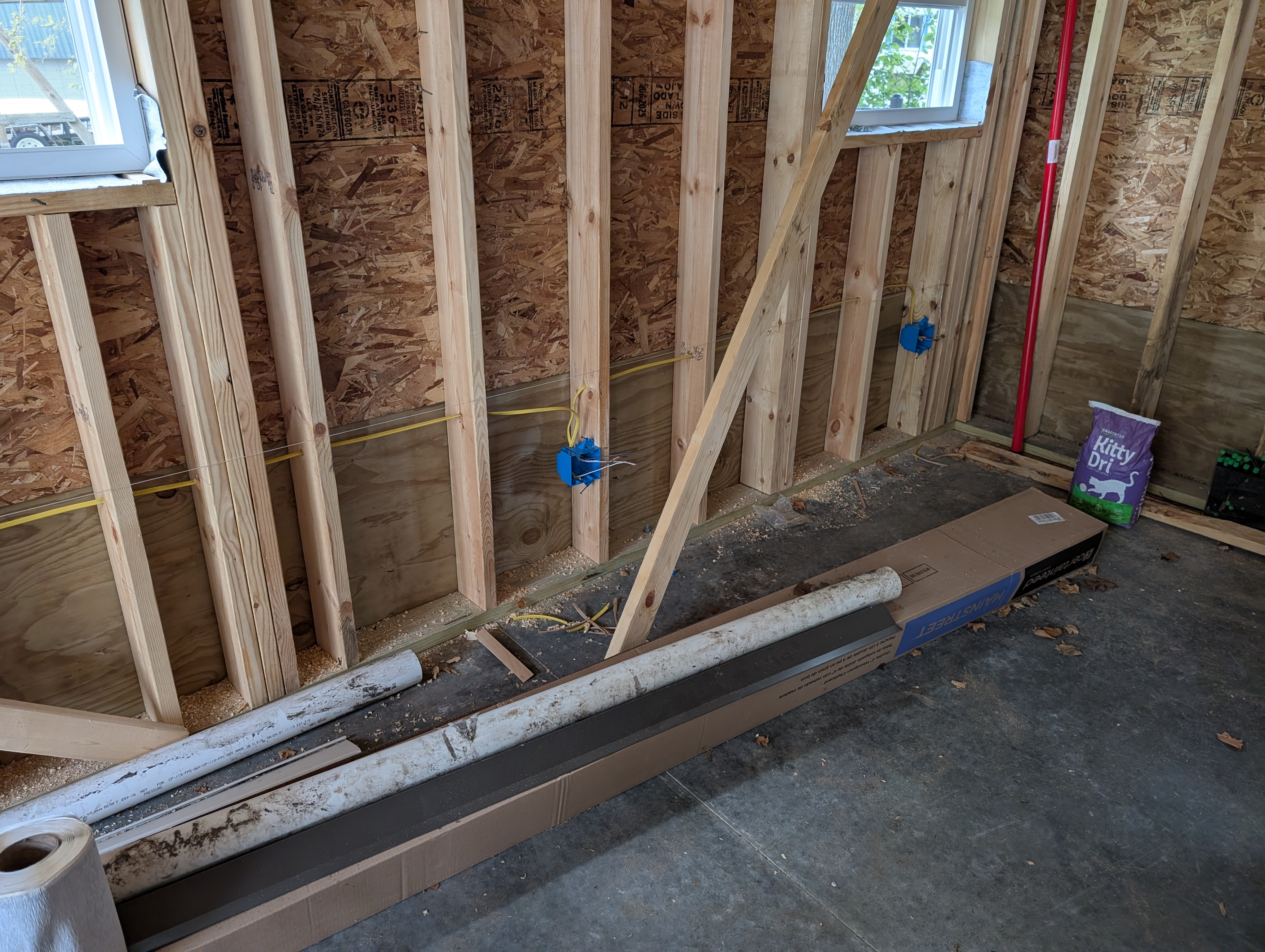 West wall view between two windows near the west/north corner; single 2-gang plastic box mounted at 16" AFF (floor-line height); PVC and trim staged on the slab below; kitty litter bag and other materials at right