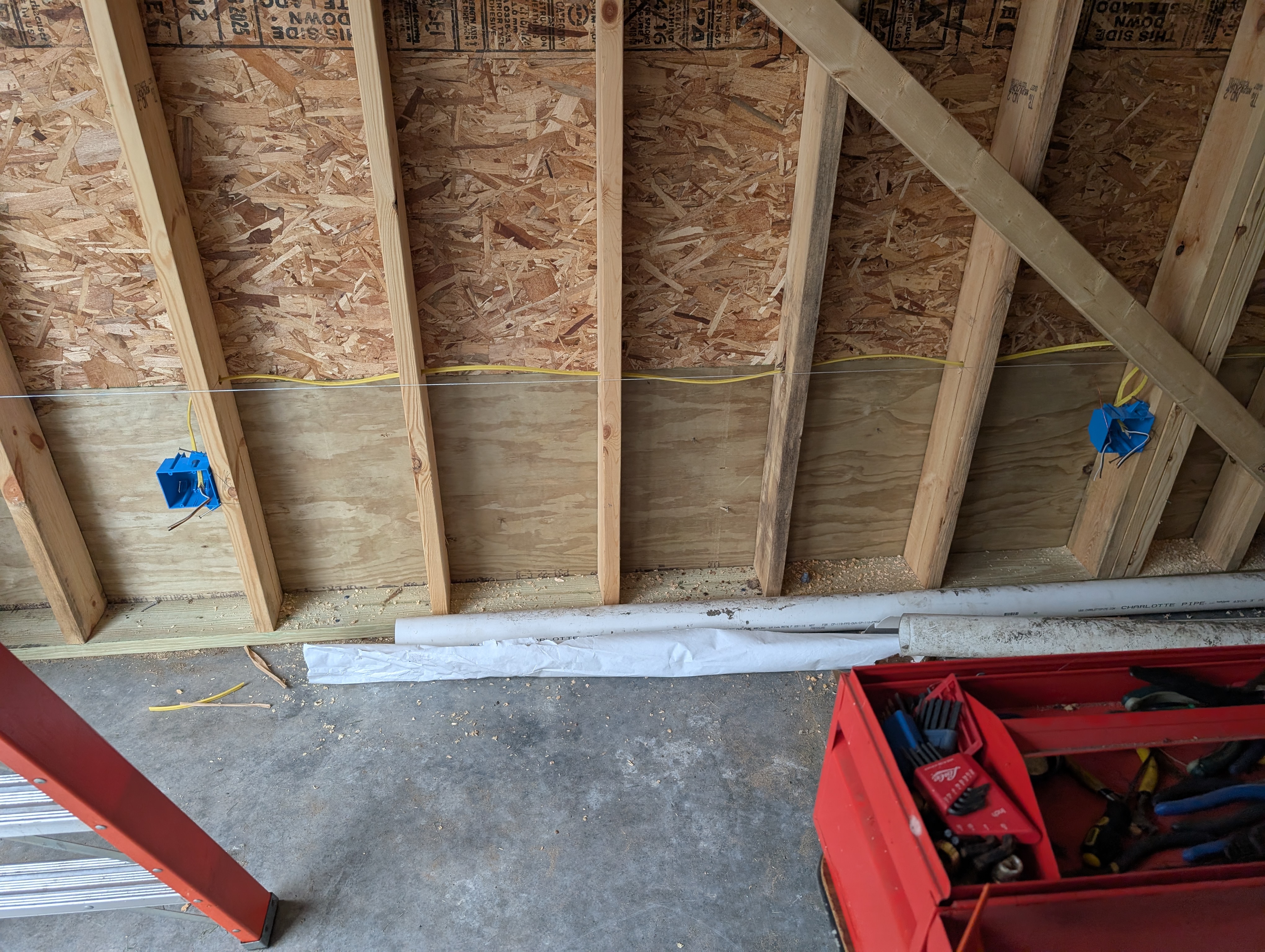 Looking down at the west wall; two blue 2-gang plastic boxes at 16" AFF on the plywood impact band with yellow romex run horizontally between them; two long PVC pipes stored on the slab running north-to-south along the wall; owner's red toolbox at lower right, ladder at lower left