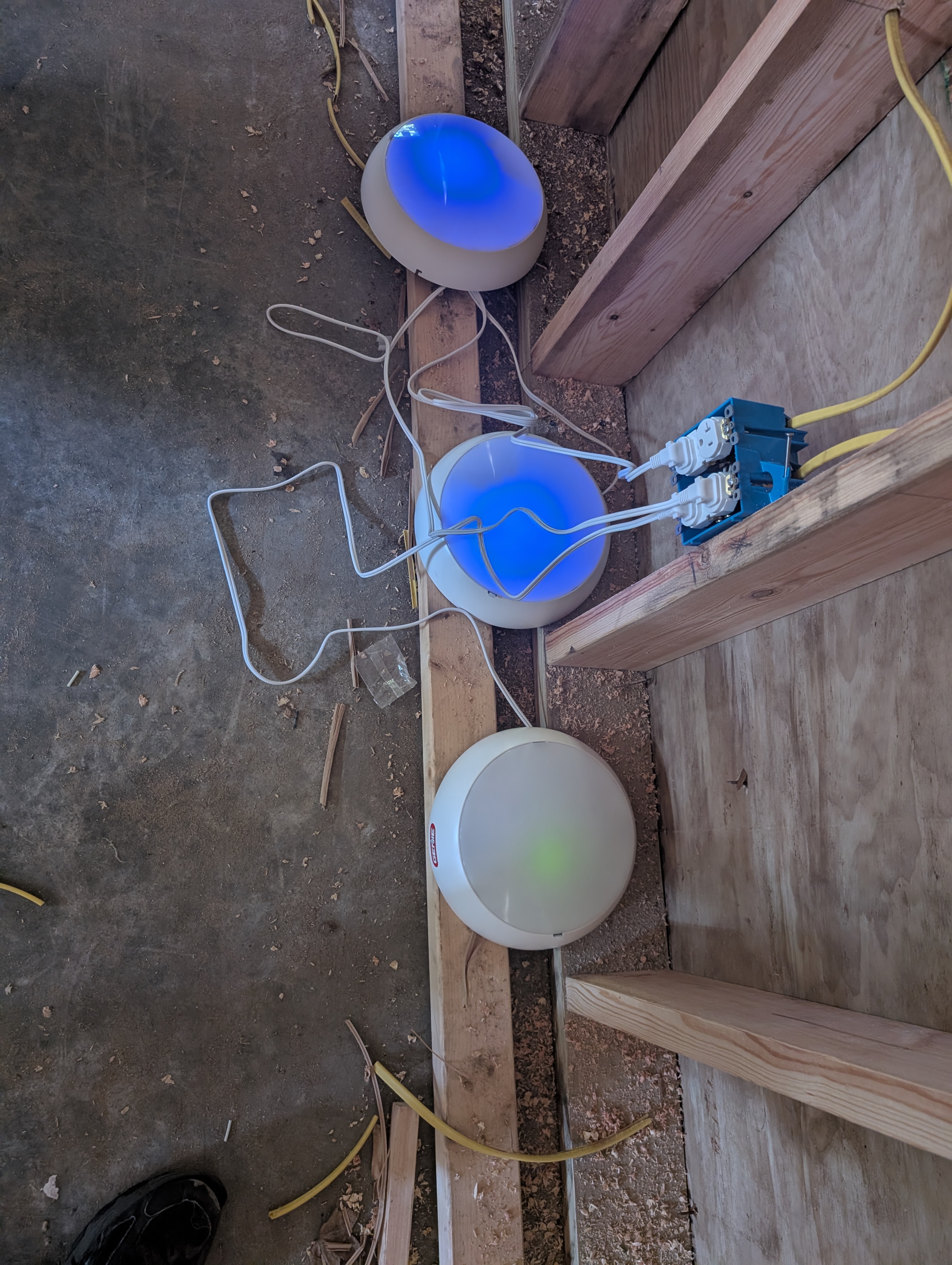 Three garage-door-opener Bluetooth safety light pucks staged on a concrete pad next to wire-nutted leads and a small junction box; two pucks glow bright blue (paired/healthy), the third glows green (interlock fault — lost Bluetooth pairing); yellow Romex visible at the edges of frame; mid-day sunlight outdoors
