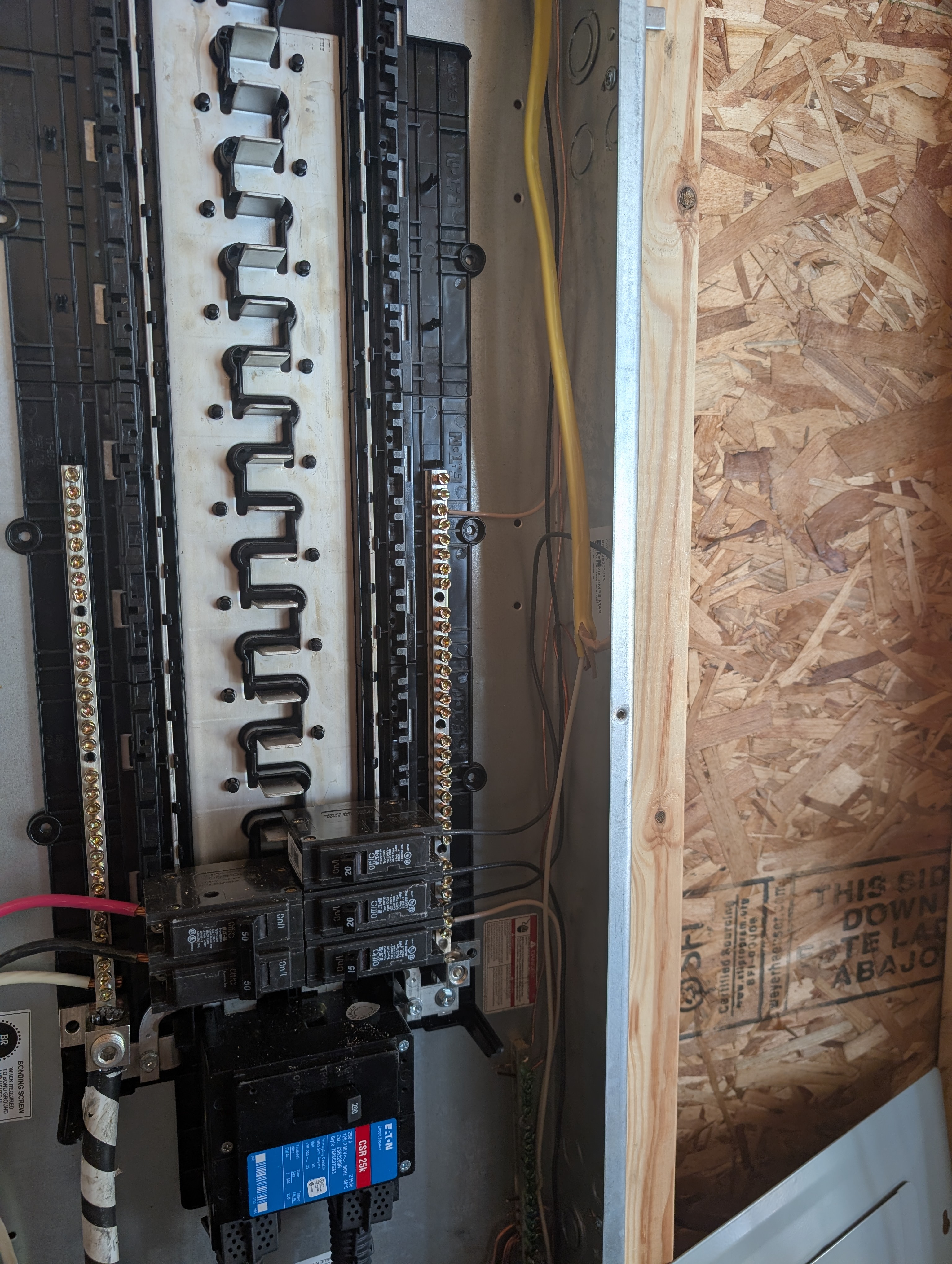 Side-angle view of the Eaton 200A panel interior with dead-front off; the bus stack visible along its full length showing the empty stab positions; new 20A BR branch breaker for the rear-wall circuit landed near the bottom of the bus stack; the 50A 2-pole generator backfeed visible at the bottom; OSB sheathing on the wall to the right