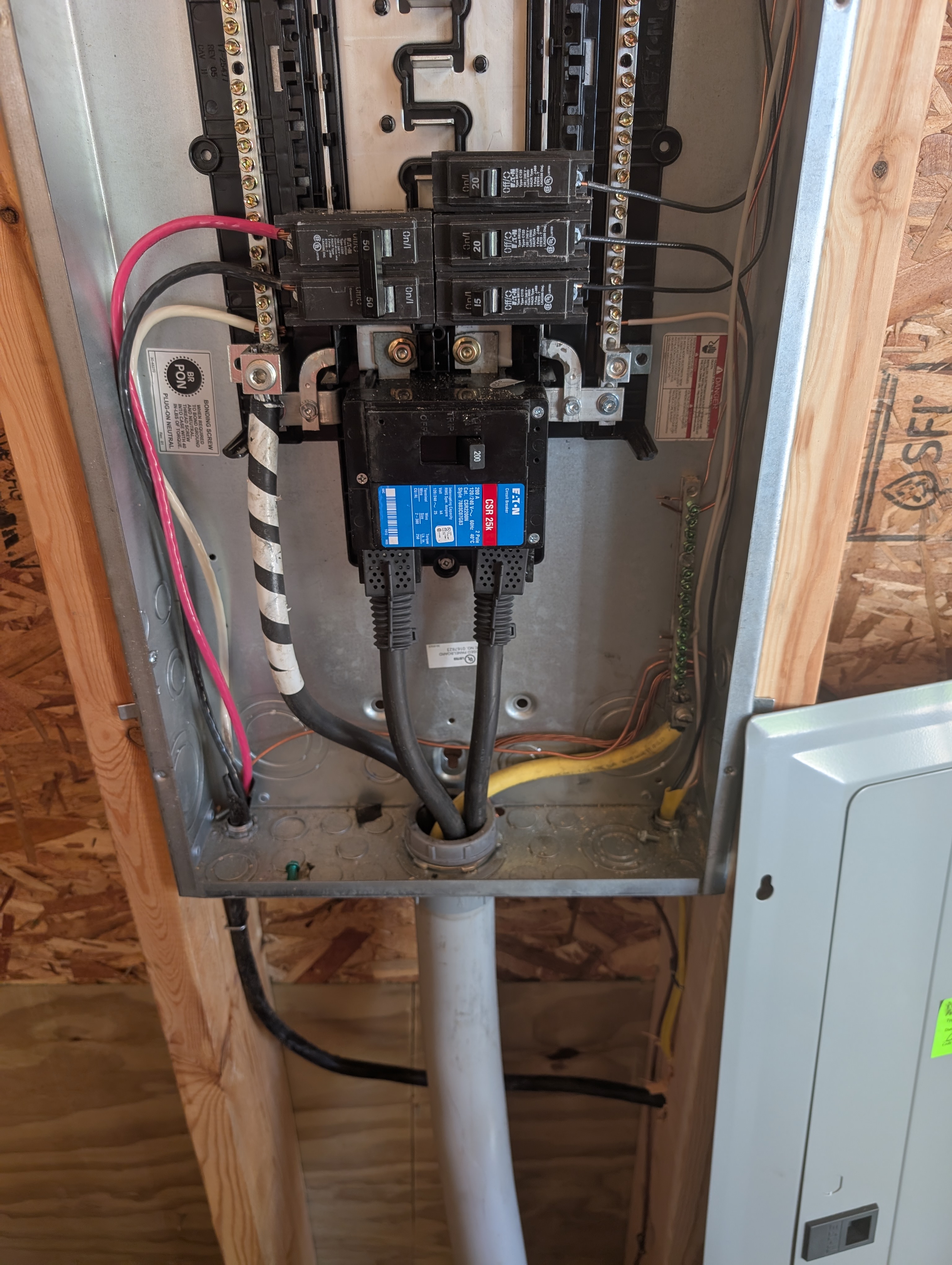Close interior view of the Eaton 200A panel with the dead-front off; 2-pole 50A breaker on the left with thick red and white feeders going off-panel to the generator inlet; black 200A main breaker block at center with thick output cables exiting the bottom; smaller 1-pole branch breakers across the bus stack including the new 20A BR for the rear-wall circuit; yellow Romex emerging through the bottom of the panel cabinet; OSB sheathing behind; gray PVC conduit and the side of an adjacent panel visible at the bottom right