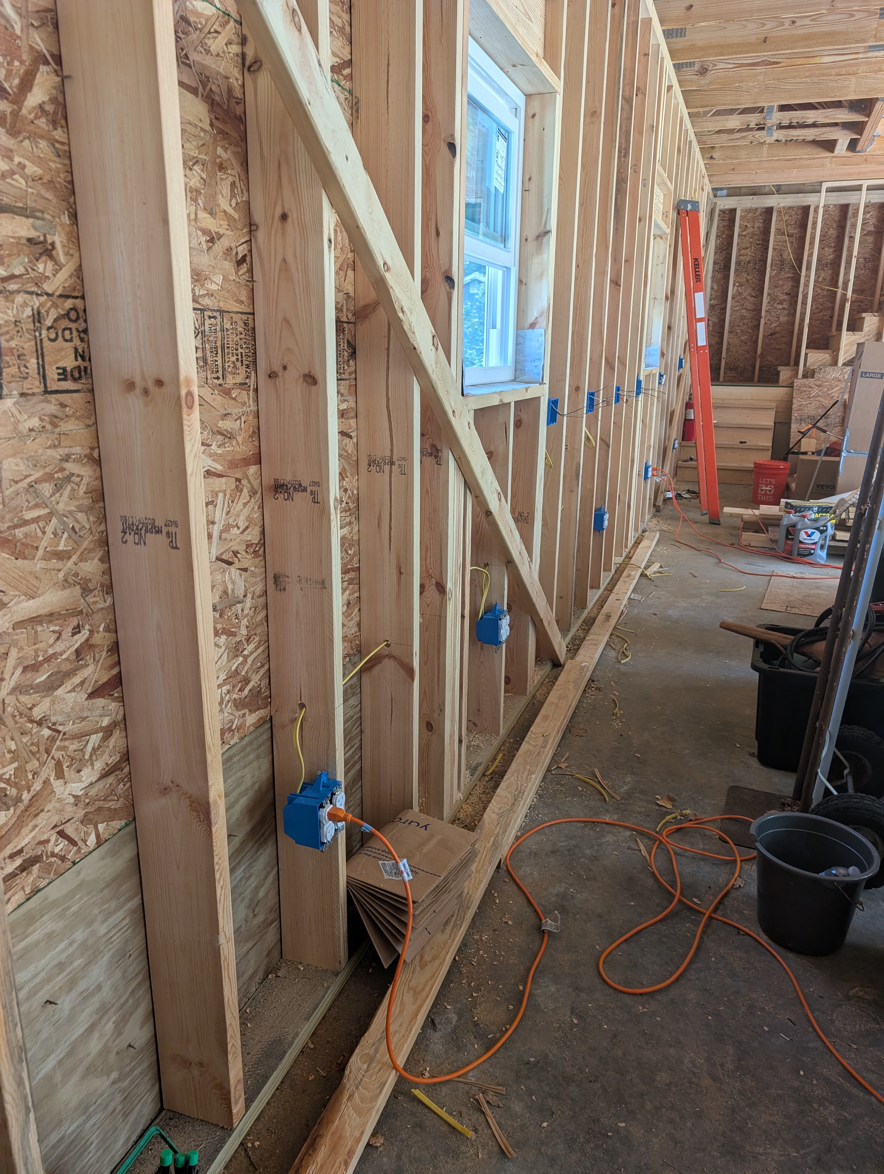Along-wall view of the north wall — multiple blue plastic boxes mounted to studs at two heights: 16" AFF floor line (lower boxes — these are on the energized rear-wall circuit) and a workbench-height (~46" AFF) line (higher 2-gang boxes — these are a separate, not-yet-energized circuit); one of the workbench-height GFCI 2-gang boxes is visible; yellow Romex chained between bays; window opening behind one of the upper boxes; raked loft stair stringer visible at upper right; orange extension cord and tools on the concrete floor