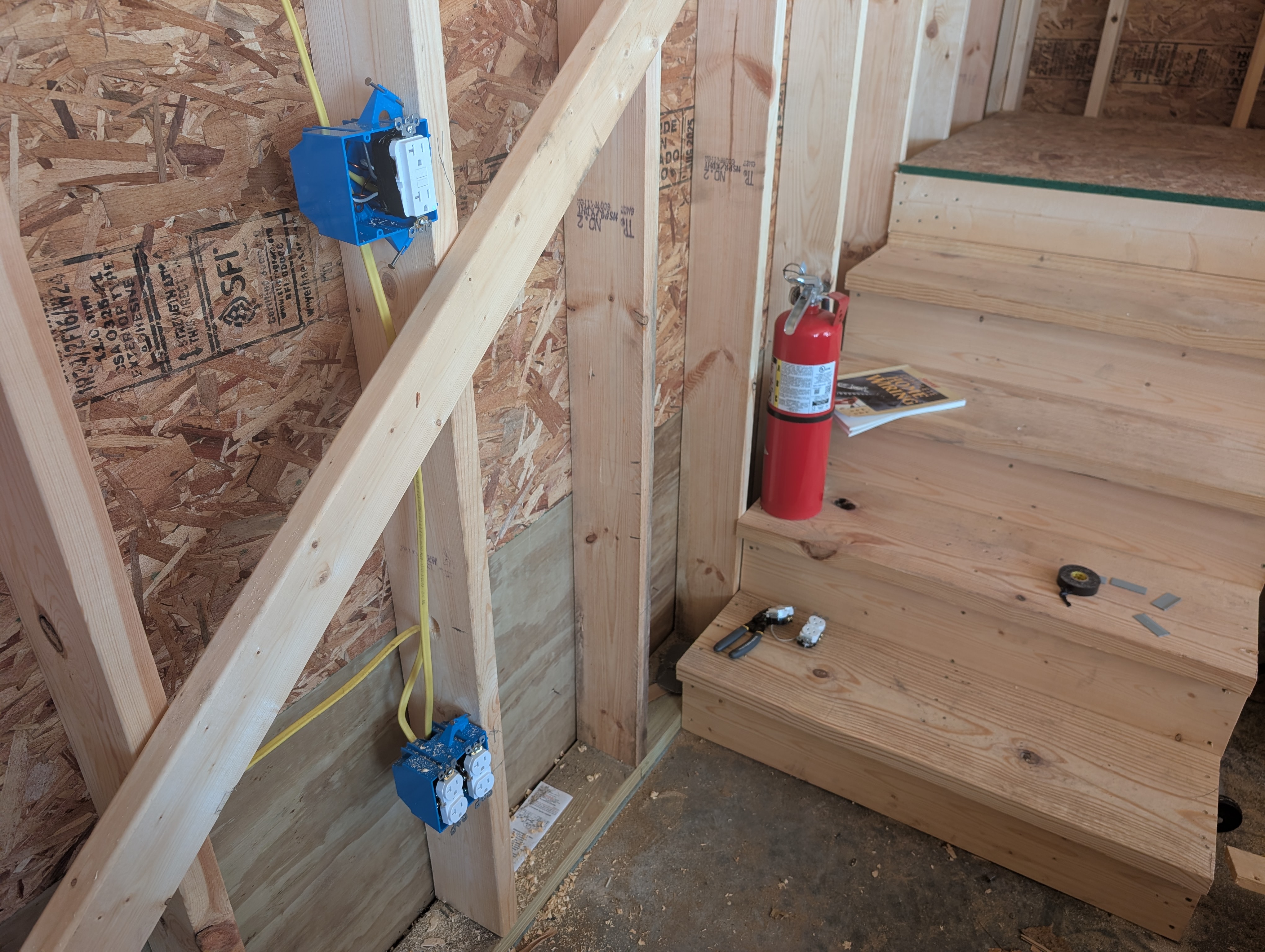 East end of the north wall near the loft stairs — upper blue 2-gang box mounted at workbench height (~46" AFF) holds the GFCI lead for the energized 16" floor-line circuit (raised above the standard perimeter height to clear the rolling tool cabinets that will sit on this section of wall); lower blue 2-gang box at 16" AFF is the first downstream box on the same circuit; yellow 12-2 NM-B Romex visible dropping from the upper box to the lower box; the bottom run of the loft stairs entering the frame from the right with a red fire extinguisher standing on the lowest tread; tape measure and Marrette wirenuts on a higher tread