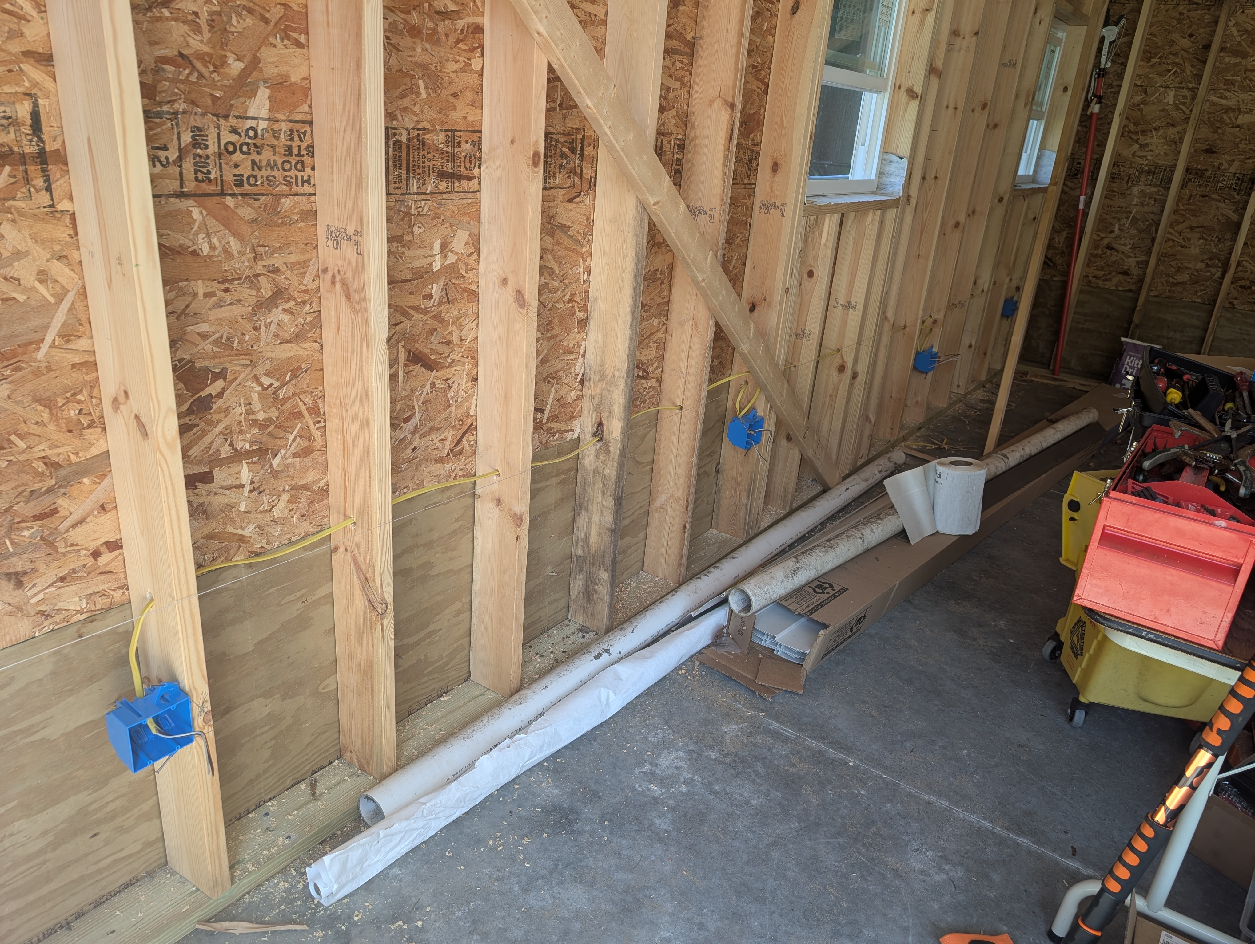 Wide angle looking along the west wall — OSB sheathing on the closer wall; a blue 1-gang plastic box mounted to a stud at 16" AFF on the left foreground; diagonal stud bracing across the frame; the rear/north wall visible in the background with a window opening and a workbench-height 2-gang box partially visible at the corner; PVC pipe and offcuts on the concrete floor; some loft framing and stairs visible at far right
