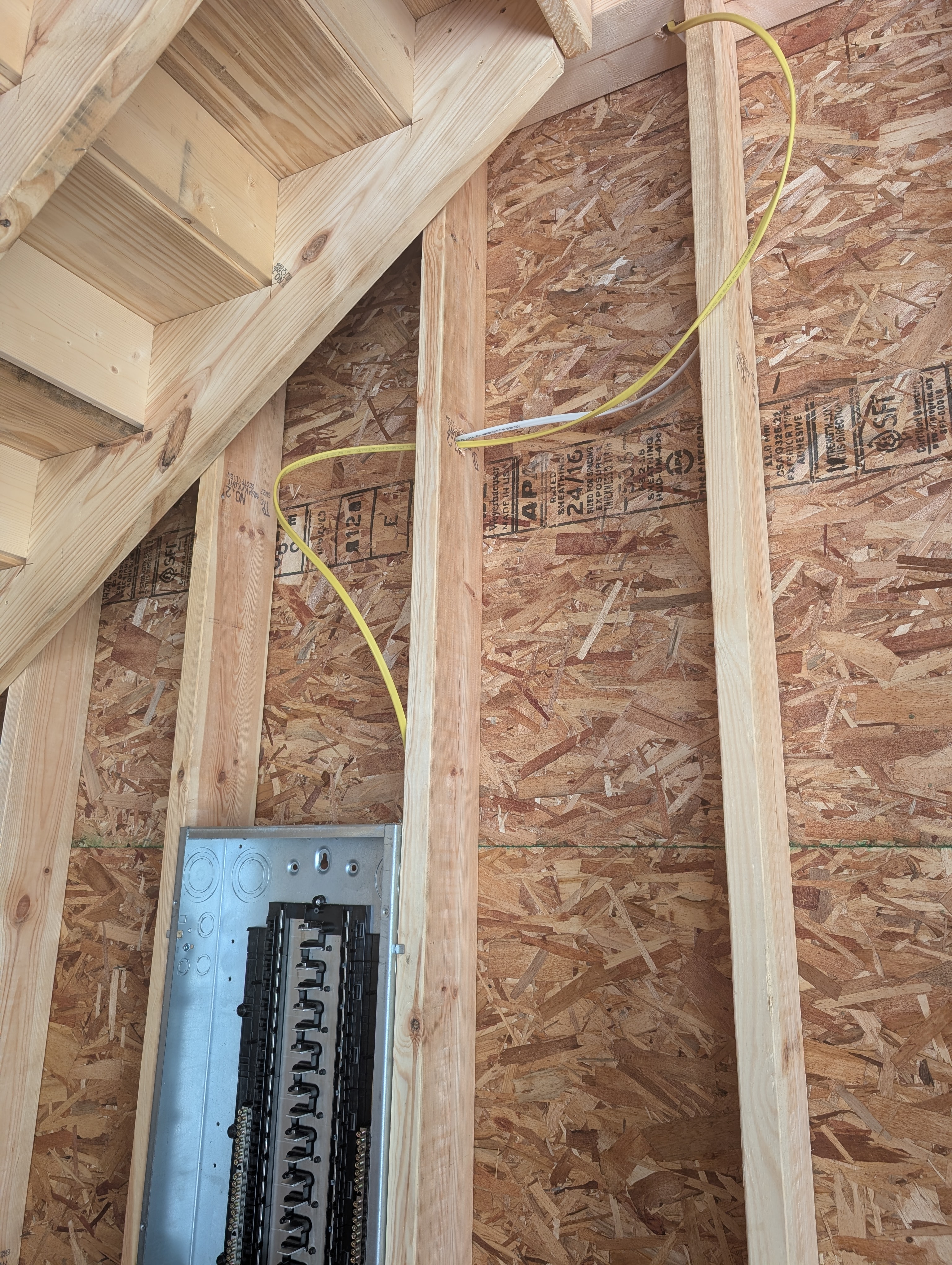 Looking up at the wall above the Eaton 200A panel; yellow 12-2 NM-B Romex emerging from the top of the panel, looping with deliberate extra length up the stud bay toward the loft floor joists / rafter space; the top edge of the panel visible at the bottom of frame; OSB sheathing on the wall behind; loft joists visible at the top of frame