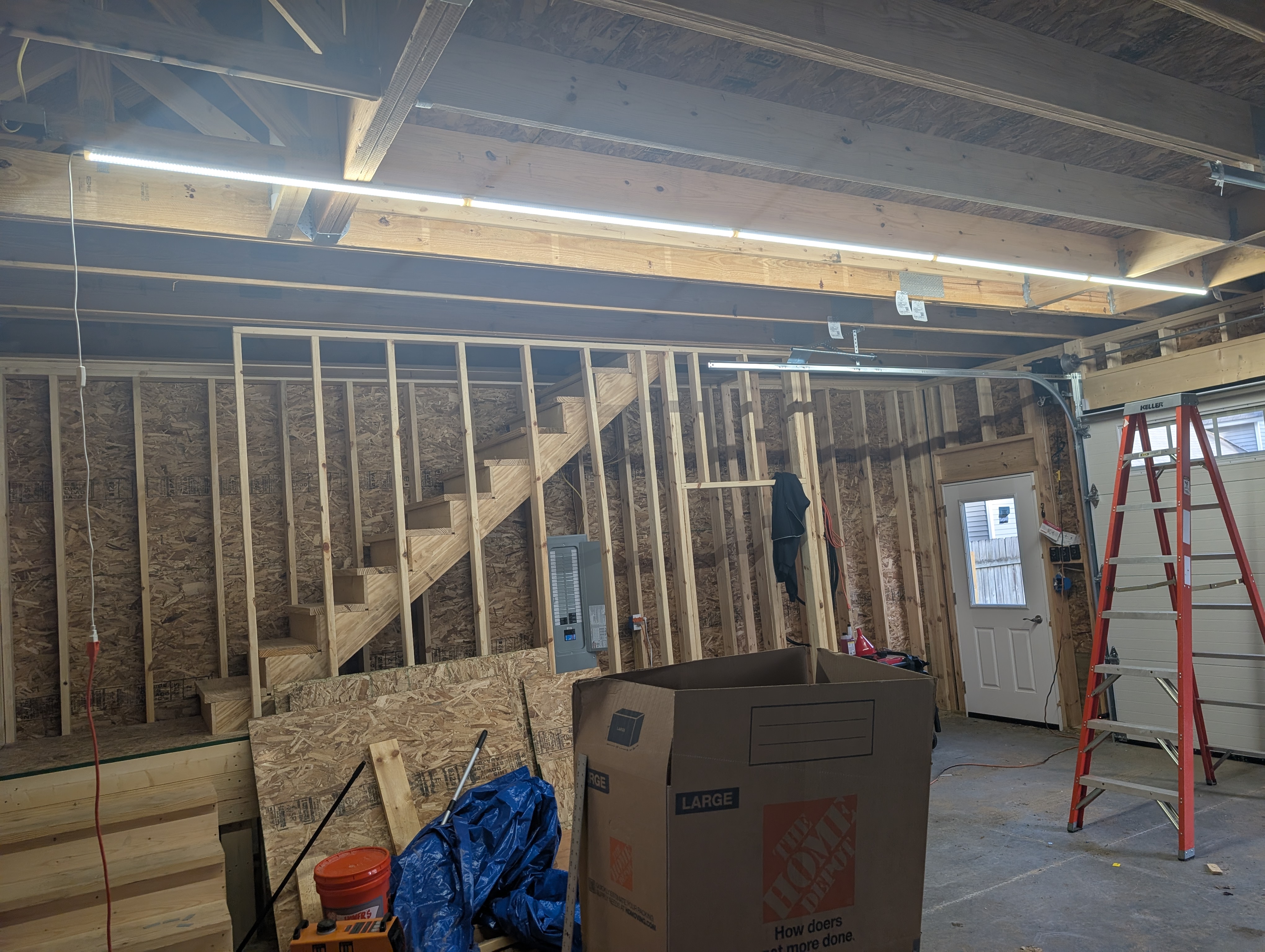 A single front-to-back run of five linked LED shop lights glowing along the garage ceiling; loft stairs and 200A panel at left, entry door and step ladder at right