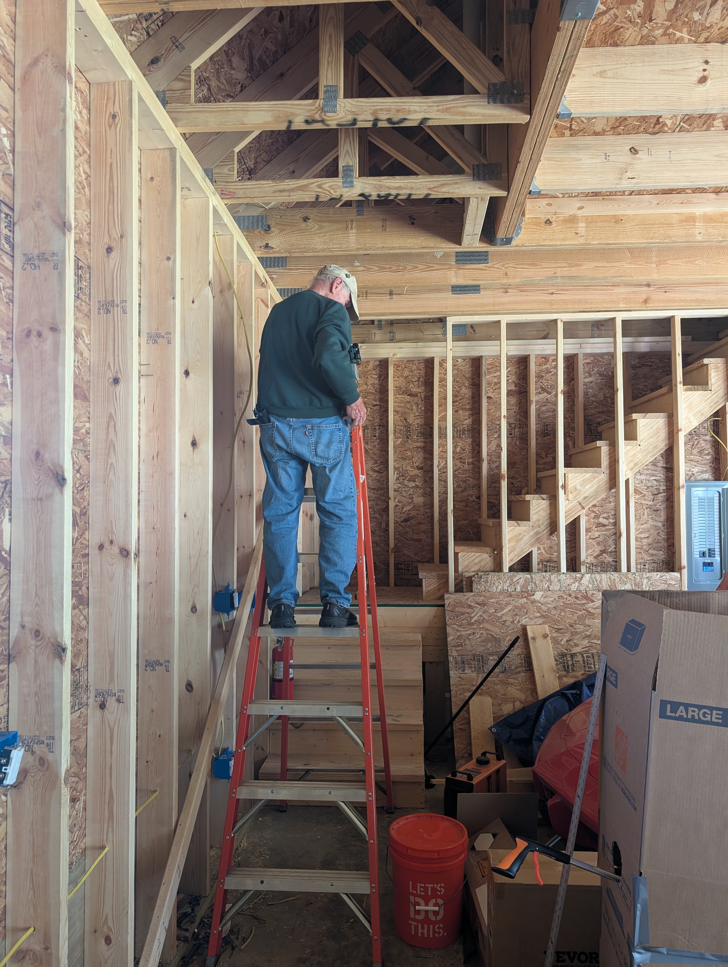 Uncle Ed on a fiberglass step ladder at the bottom of the loft stairs, reaching up to run cable to the ceiling-light boxes near the stairwell switch location; the garage 200A panel is on the wall at right