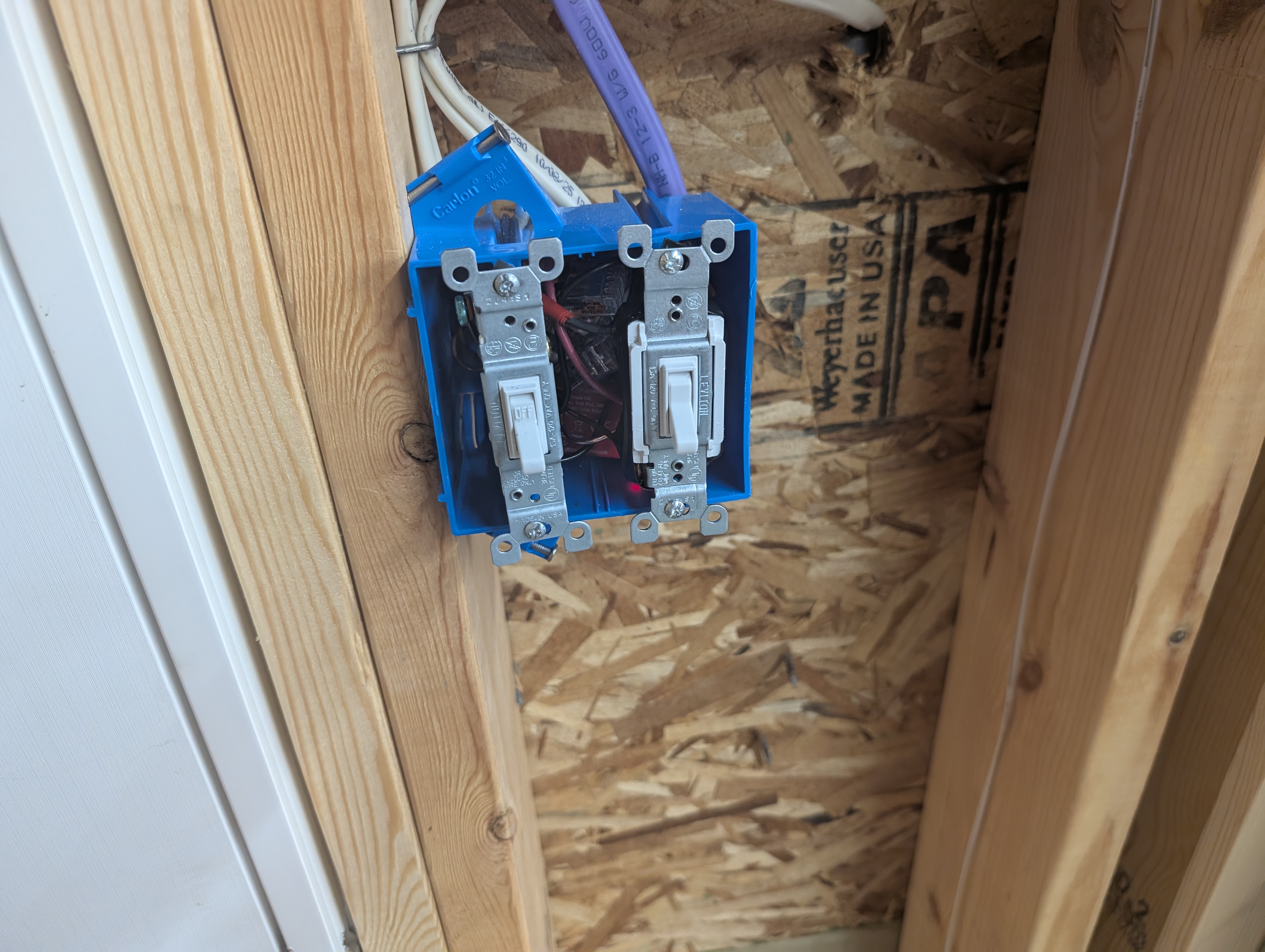 Entry/service-door 2-gang switch box with both toggles installed: the existing exterior-light switch and the new garage-floor-light 3-way, with the 12/3 landed and the red traveler visible