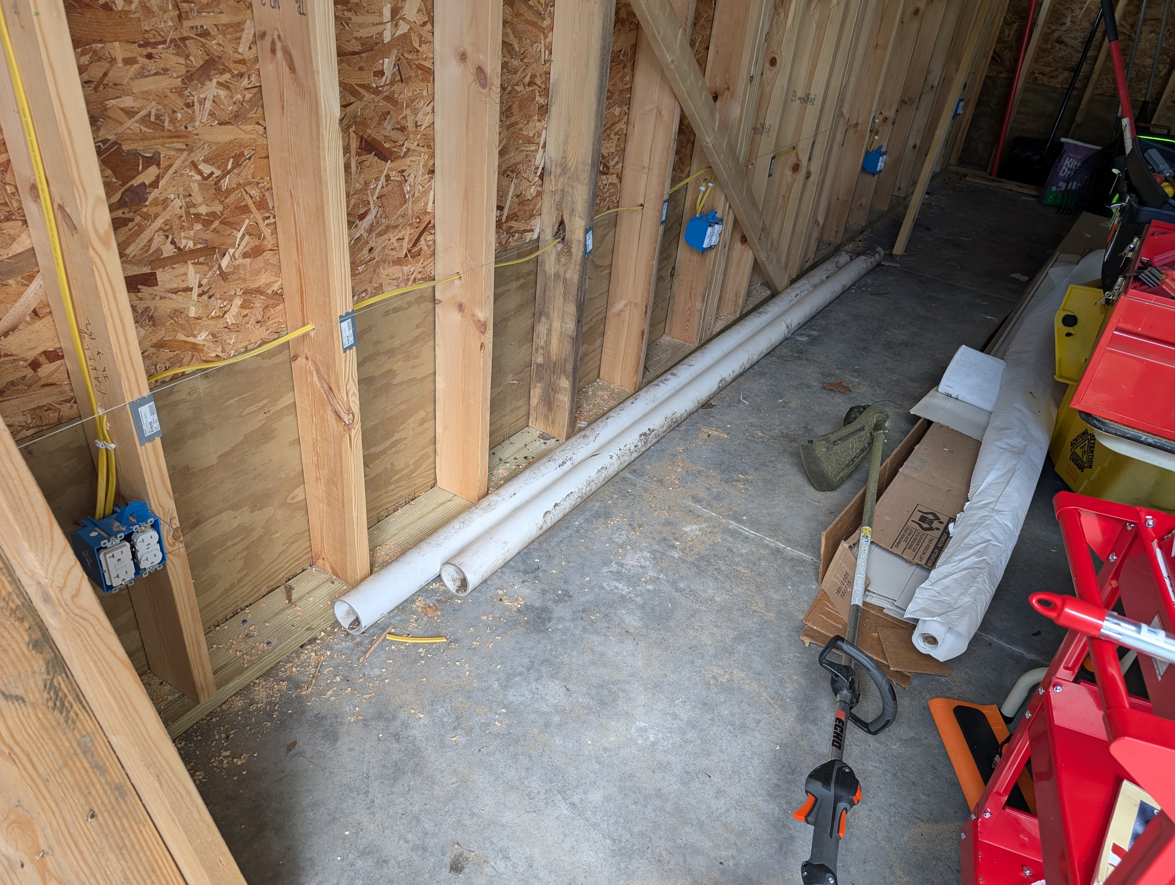 West wall electrical rough-in viewed from the garage door opening, showing the dual-gang GFCI feed box, the NM-B run down the wall, and nail plates protecting the cable