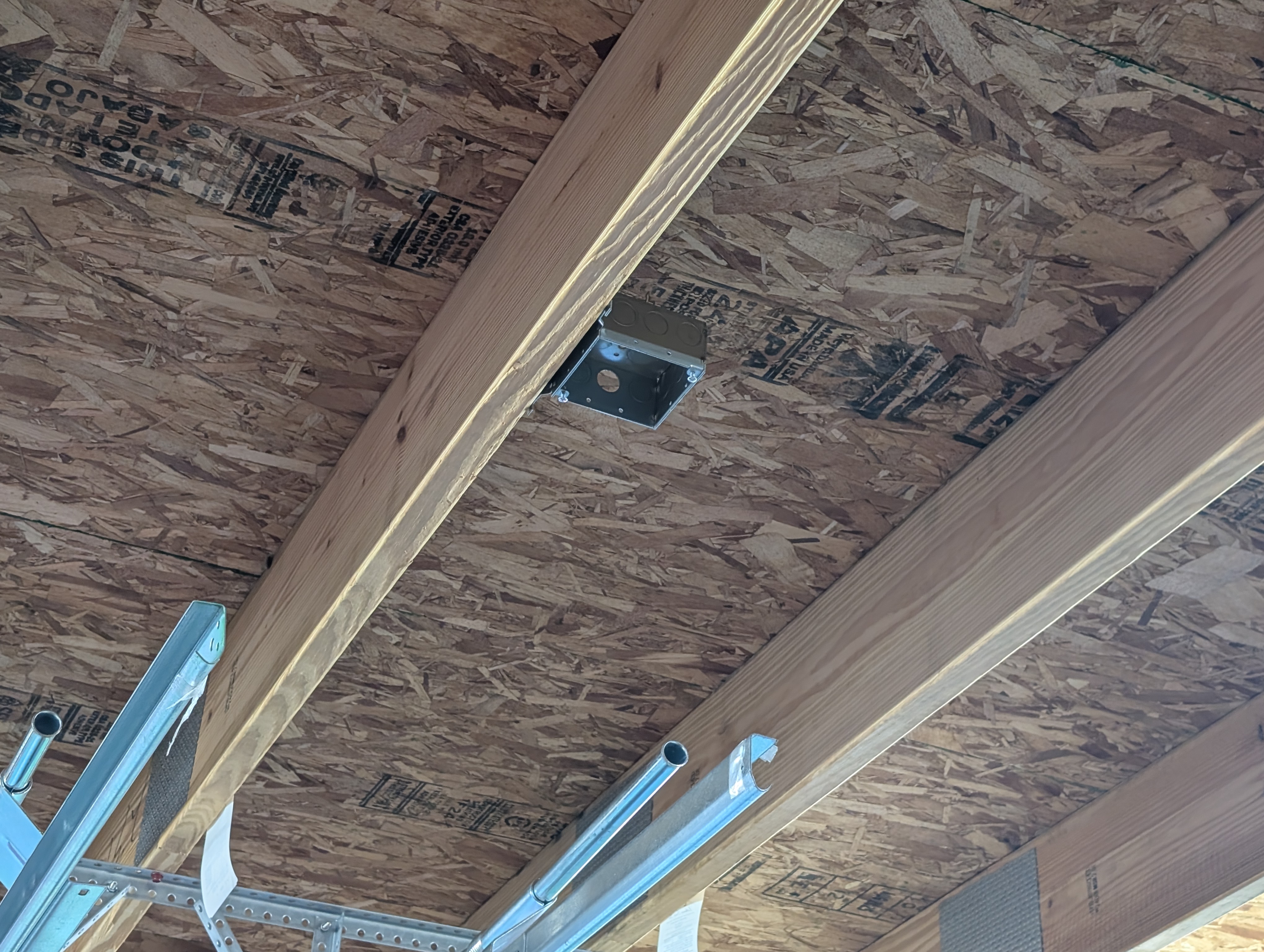 Metal ceiling box mounted flush to the OSB roof sheathing alongside a ceiling beam, near the center of the garage between garage-door bays; a garage-door opener rail runs below