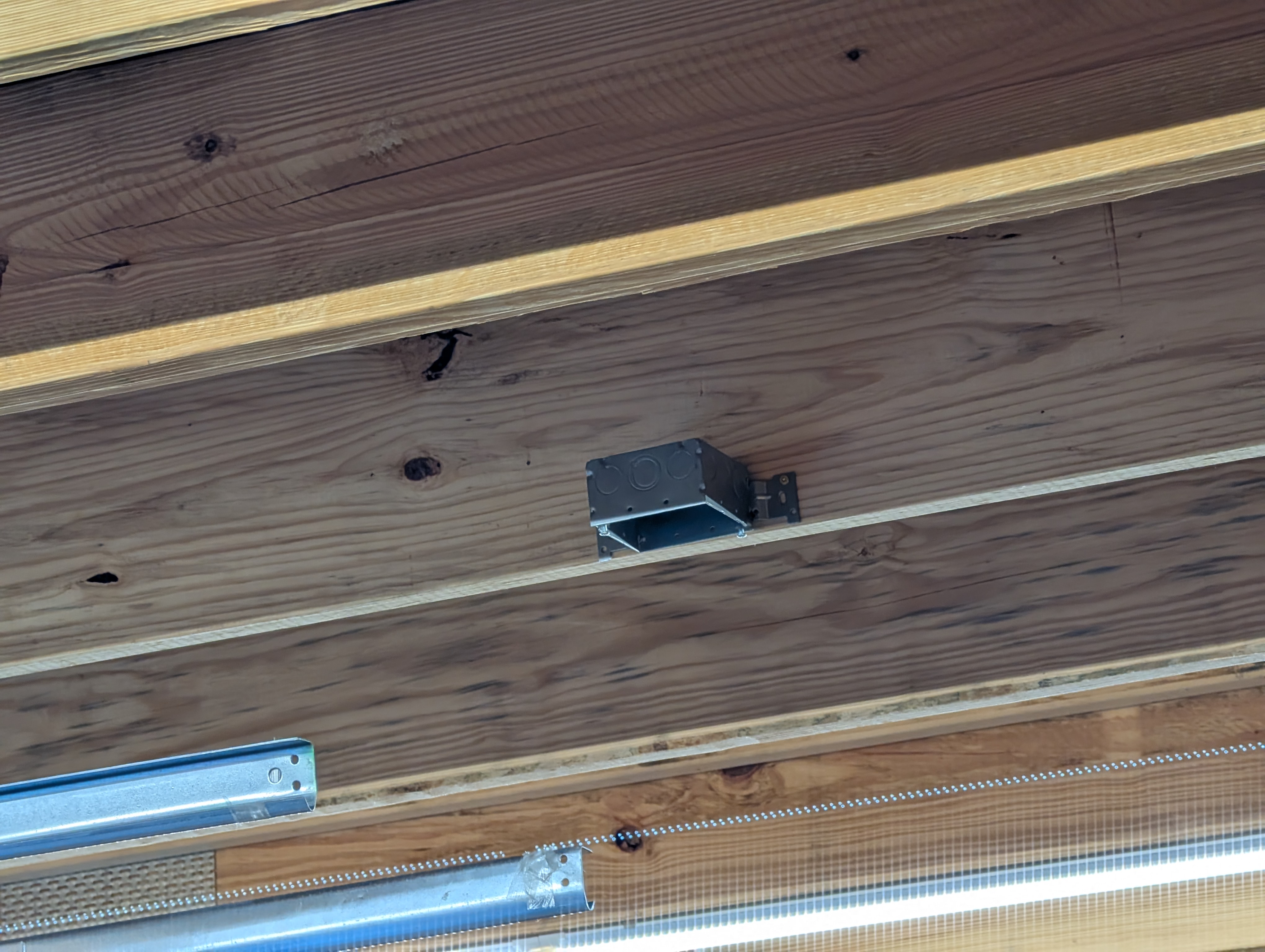 Single-gang metal ceiling box mounted to a ceiling purlin against the wood sheathing, near the center of the garage between garage-door bays; opener rails visible below