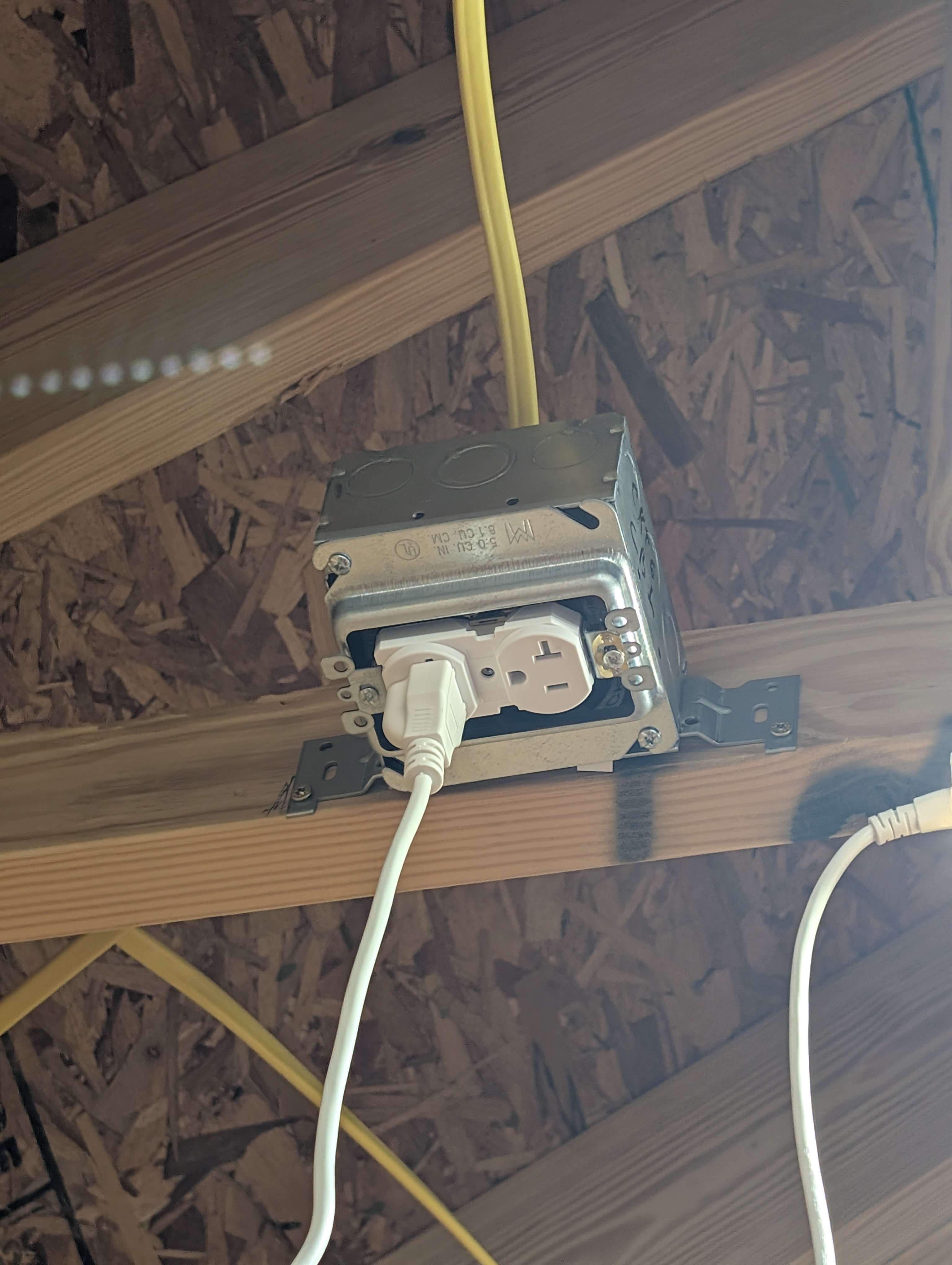 4-inch square ceiling box on a bar hanger between two truss bottom chords, fitted with a mud ring and a 20A tamper-resistant duplex receptacle; a white Barrina LED power cord is plugged in and a yellow 12/2 NM-B daisy-chain enters the top of the box