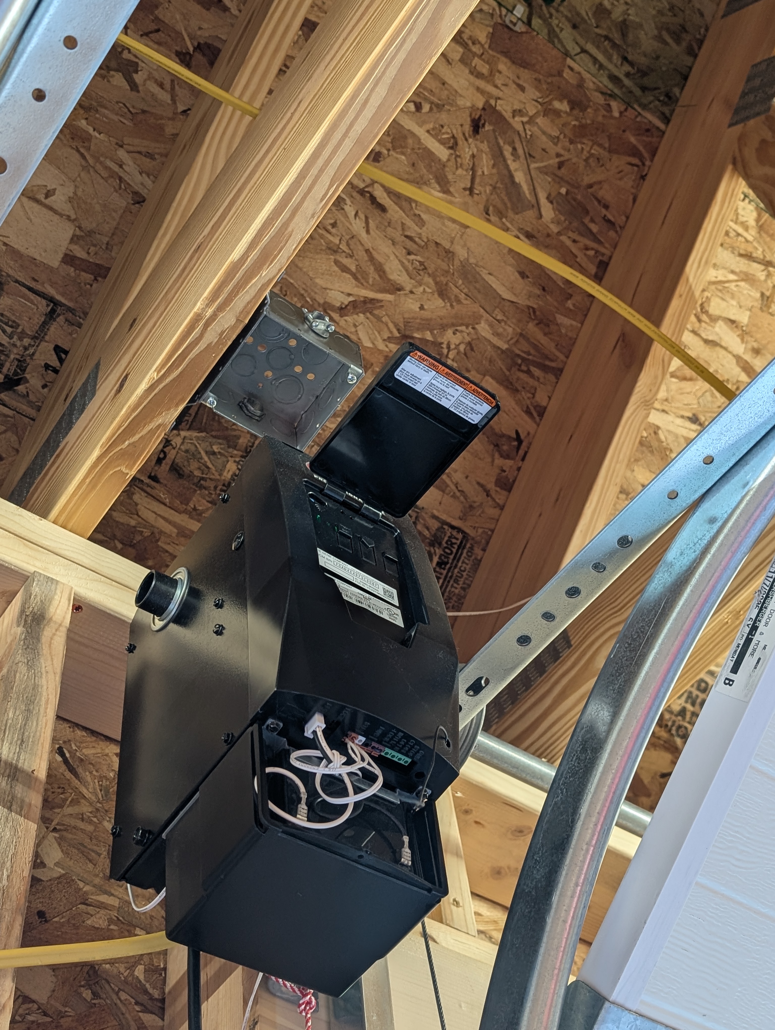 Genie garage-door opener head with a metal ceiling box mounted on the framing above it; opener rail and OSB sheathing visible