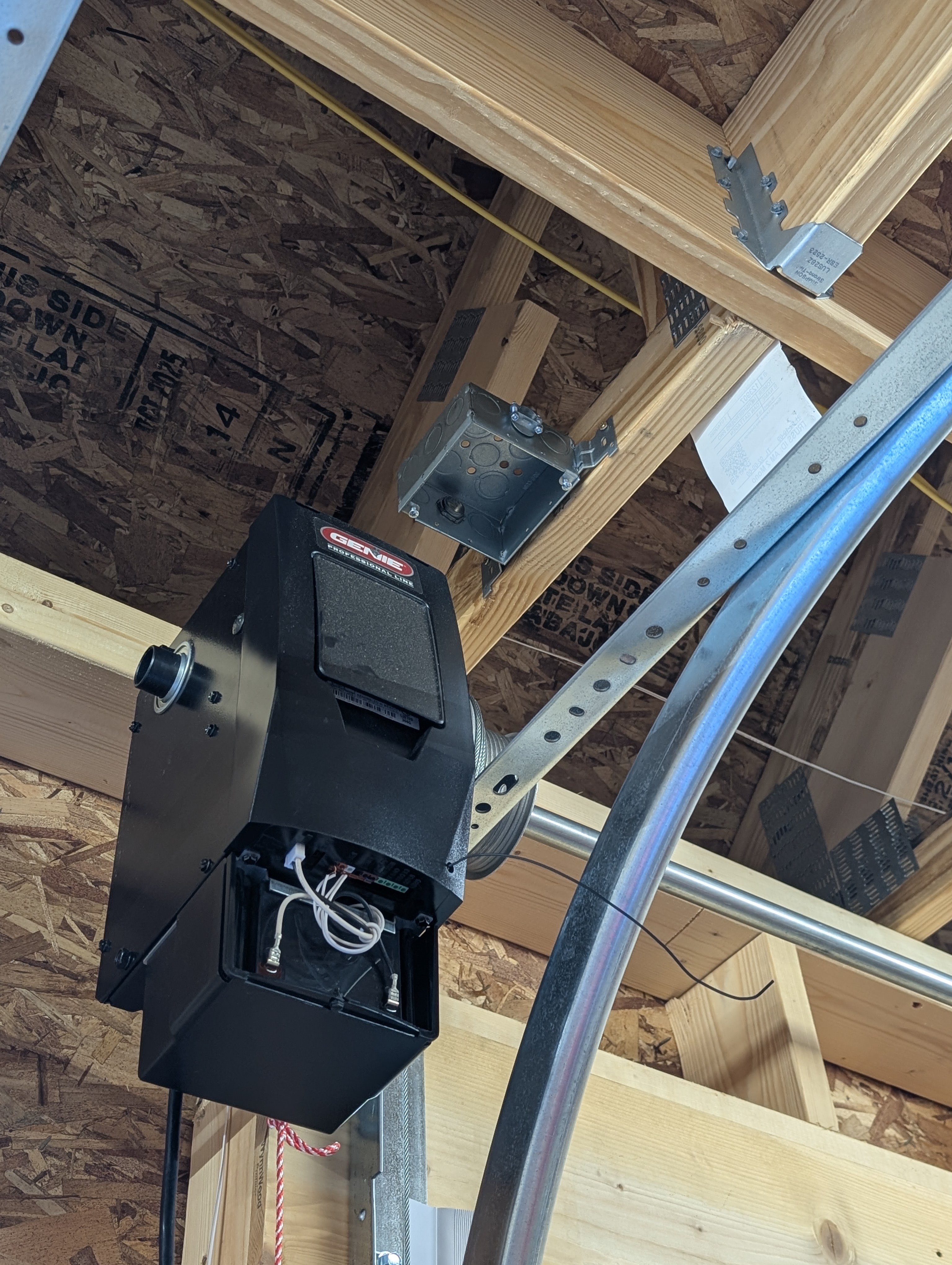 Genie garage-door opener head with a metal ceiling box mounted on the framing above it; opener rail, conduit, and OSB sheathing visible
