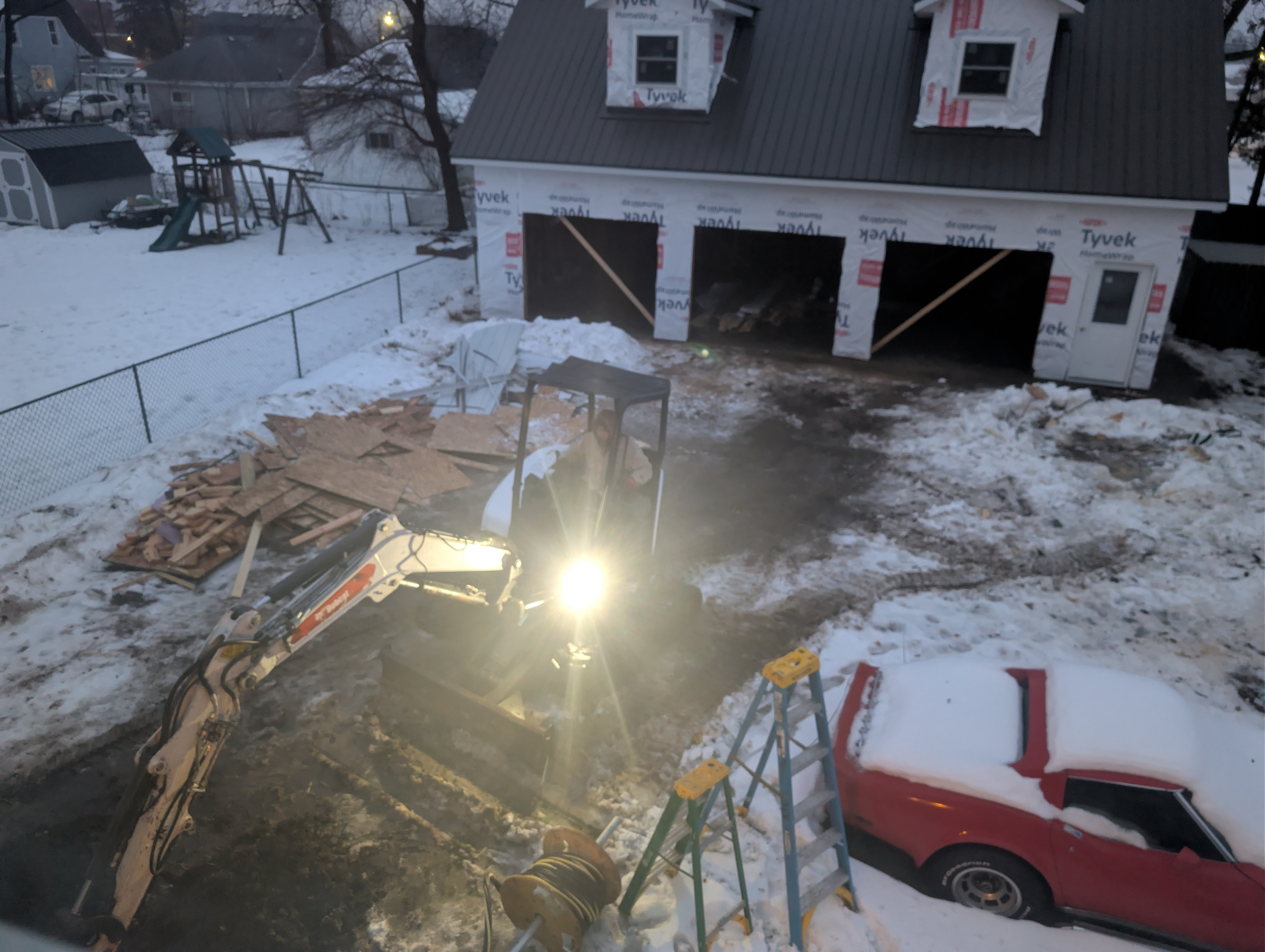 Backhoe attempting to dig in frozen ground with garage in background at dusk